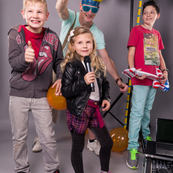 Der DJ und drei Kinder, die ein Mikrofon und eine Luftgitarre halten. Im Hintergrund sind Luftballons.